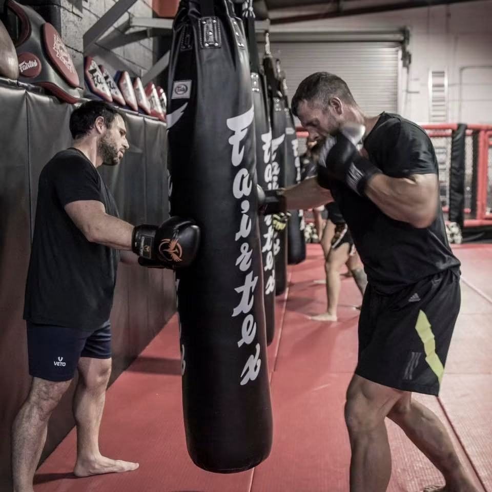 Member throwing a right hand into a heavy bag while a coach holds it steady at Chau Kaizen