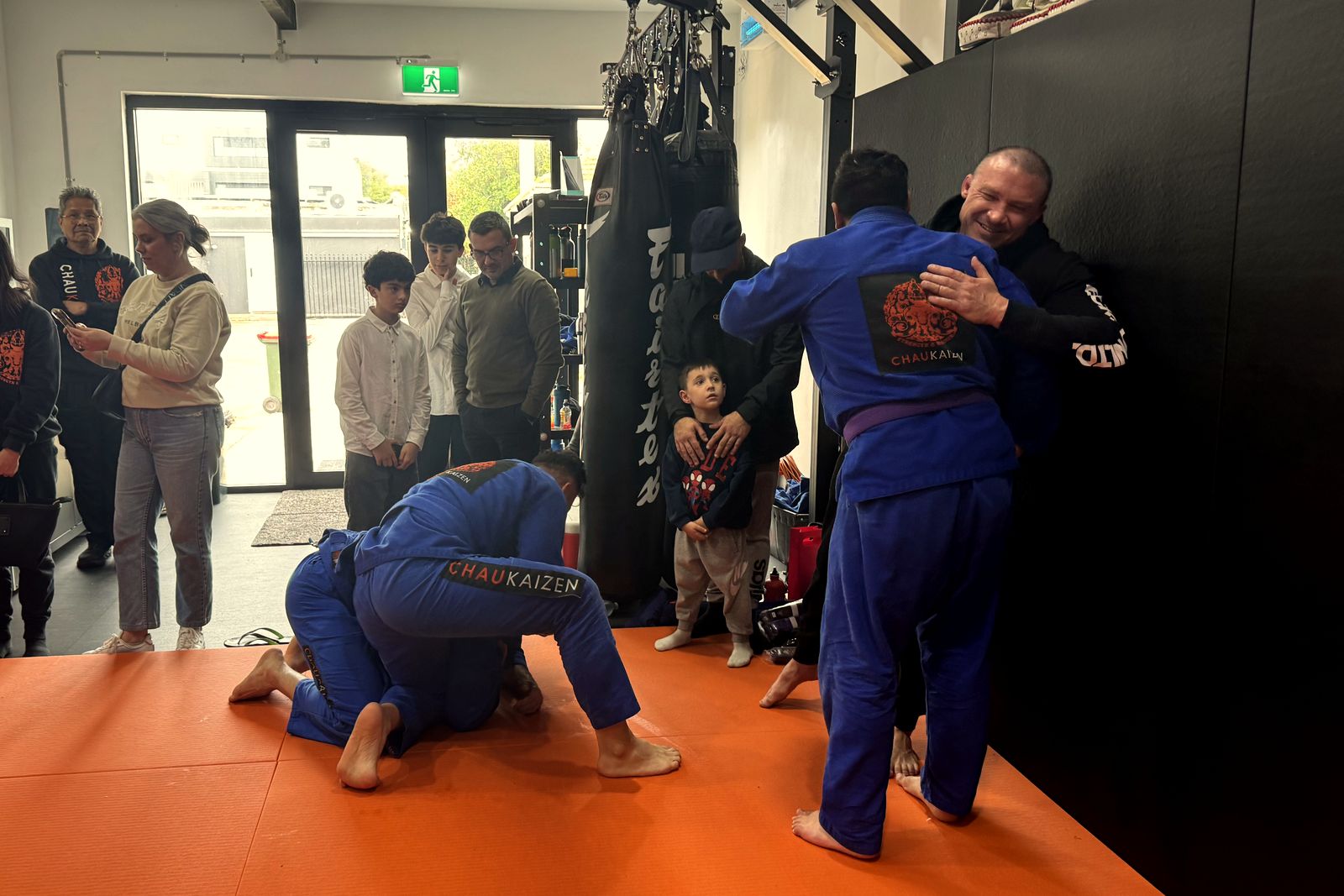 A coach hugs a member in a blue gi and purple belt while two members roll on the mats and families watch, at Chau Kaizen in Prospect