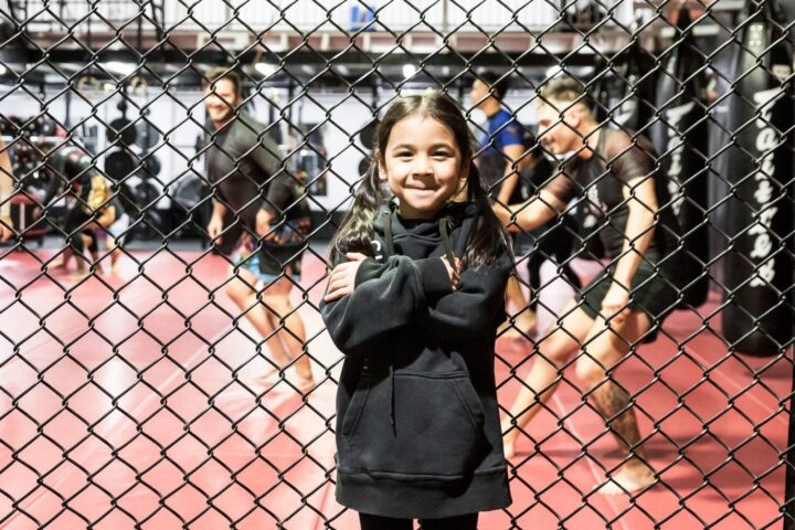Head coach Suasday Chau's daughter smiles with her arms crossed at the cage fence while adults train behind her, at Chau Kaizen's old facility